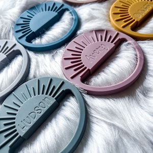 May include: Five colorful silicone teethers shaped like suns with names inscribed on them. The teethers are on a white fuzzy surface.
