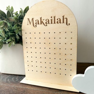 May include: Wooden earring holder with a rounded top and a stand. The holder has a grid of holes for storing earrings and the name "Makailah" is engraved at the top.