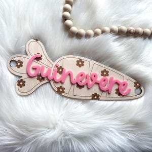 May include: A wooden carrot-shaped sign with a pink floral pattern and the name "Guinevere" written in pink letters.