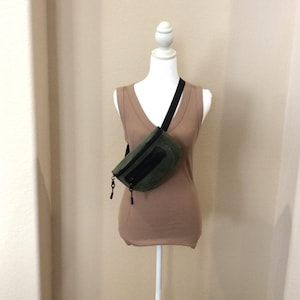 May include: An olive green fanny pack with a black zipper closure and a black adjustable strap, worn across the chest over a tan sleeveless top on a white mannequin. The bag features a textured fabric surface and black zipper pulls.