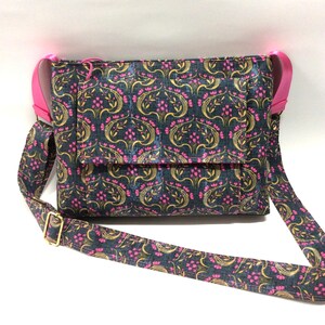 May include: A crossbody bag with a floral pattern in navy, gold, and pink. The bag has a flap closure and a pink strap. The adjustable strap is patterned with the same floral design and has a gold-colored buckle.