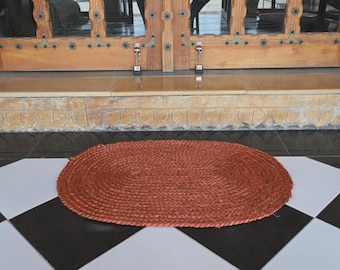 Könnte beinhalten: Ovaler, gewebter Jute-Teppich in einem warmen, rötlich-braunen Farbton. Der Teppich liegt auf einem schwarz-weiß karierten Fliesenboden vor einer Holztür mit dekorativen Paneelen. Die Textur und Farbe des Teppichs verleihen dem Eingangsbereich ein natürliches Element.
