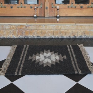 Peut inclure: Un tapis rectangulaire gris foncé avec un motif géométrique central en nuances de gris et de blanc. Deux rayures verticales longent les côtés. Le tapis a une bordure effilochée et est posé sur un sol à carreaux noir et blanc, devant une porte en bois.