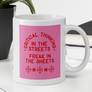 May include: White ceramic mug with a pink rectangle featuring the text "CRITICAL THINKING IN THE STREETS FREAK IN THE SHEETS" in red. The mug is on a white surface with a keyboard, a plant, and a book in the background.