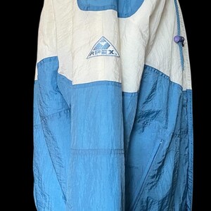 K&ouml;nnte beinhalten: Eine blaue, wei&szlig;e und hellblaue Apex-Jacke mit Kapuze. Die Jacke hat einen Rei&szlig;verschluss und ein kleines Logo auf der Brust. Die Jacke besteht aus einem leichten, geknitterten Material.