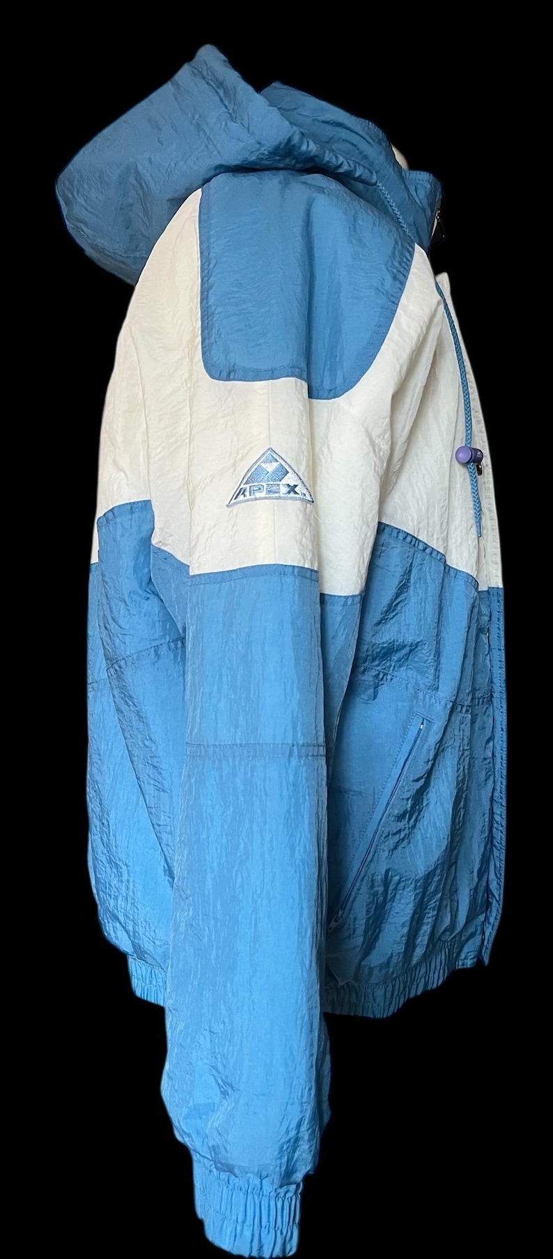 K&ouml;nnte beinhalten: Eine blaue, wei&szlig;e und hellblaue Apex-Jacke mit Kapuze. Die Jacke hat einen Rei&szlig;verschluss und ein kleines Logo auf der Brust. Die Jacke besteht aus einem leichten, geknitterten Material.