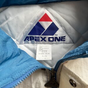 K&ouml;nnte beinhalten: Nahaufnahme einer Vintage Apex One Jacke. Die Jacke hat ein wei&szlig;es und hellblaues Farbschema mit Rei&szlig;verschluss. Das Apex One-Logo ist auf dem Etikett und dem Rei&szlig;verschlussanh&auml;nger zu sehen. Die Jacke ist in der Gr&ouml;&szlig;e XL/TG.