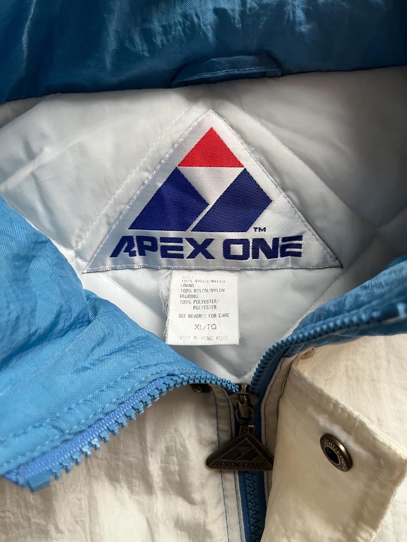 K&ouml;nnte beinhalten: Nahaufnahme einer Vintage Apex One Jacke. Die Jacke hat ein wei&szlig;es und hellblaues Farbschema mit Rei&szlig;verschluss. Das Apex One-Logo ist auf dem Etikett und dem Rei&szlig;verschlussanh&auml;nger zu sehen. Die Jacke ist in der Gr&ouml;&szlig;e XL/TG.