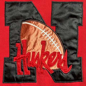 May include: Red sweatshirt with a black felt applique of the letter 'N' with a brown football and the word 'Huskers' in red.