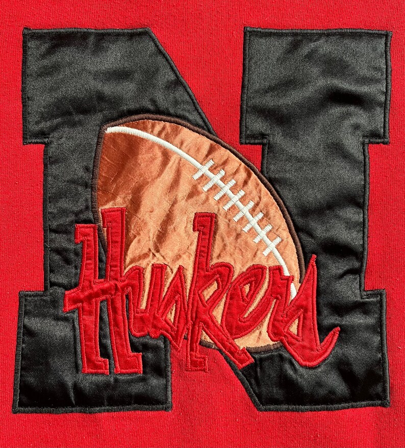 May include: Red sweatshirt with a black felt applique of the letter 'N' with a brown football and the word 'Huskers' in red.