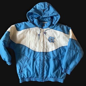 K&ouml;nnte beinhalten: Eine hellblaue, wei&szlig;e und blaue Kapuzenjacke mit Rei&szlig;verschluss. Die Jacke hat ein Logo auf der Brust und eine Kapuze mit Kordelzug. Die Jacke ist aus leichtem Material gefertigt und hat eine Vintage-&Auml;sthetik.