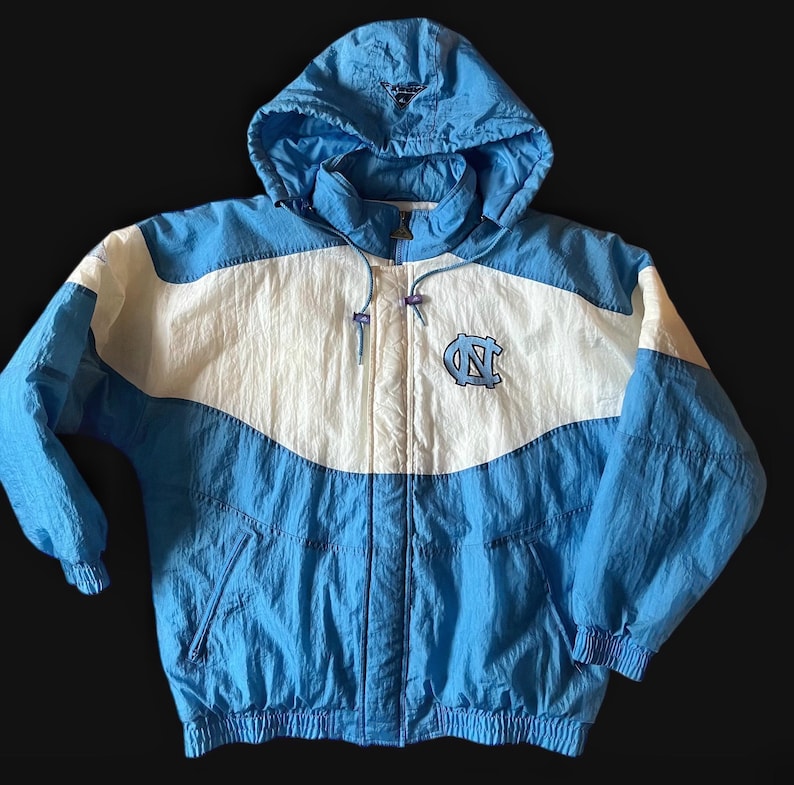 K&ouml;nnte beinhalten: Eine hellblaue, wei&szlig;e und blaue Kapuzenjacke mit Rei&szlig;verschluss. Die Jacke hat ein Logo auf der Brust und eine Kapuze mit Kordelzug. Die Jacke ist aus leichtem Material gefertigt und hat eine Vintage-&Auml;sthetik.