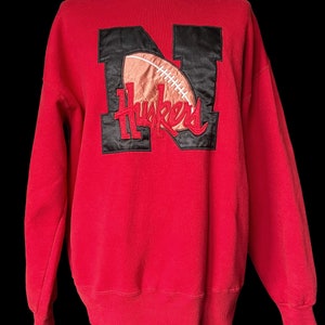May include: Red sweatshirt with a black and white embroidered "N" with a football and the word "Huskers" underneath.