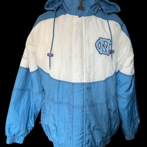 K&ouml;nnte beinhalten: Eine Kapuzenjacke in Hellblau und Wei&szlig; mit einem Colorblock-Design. Die Jacke hat einen Rei&szlig;verschluss und ein Logo auf der Brust. Die Jacke besteht aus einem gl&auml;nzenden, m&ouml;glicherweise Nylon-Material.