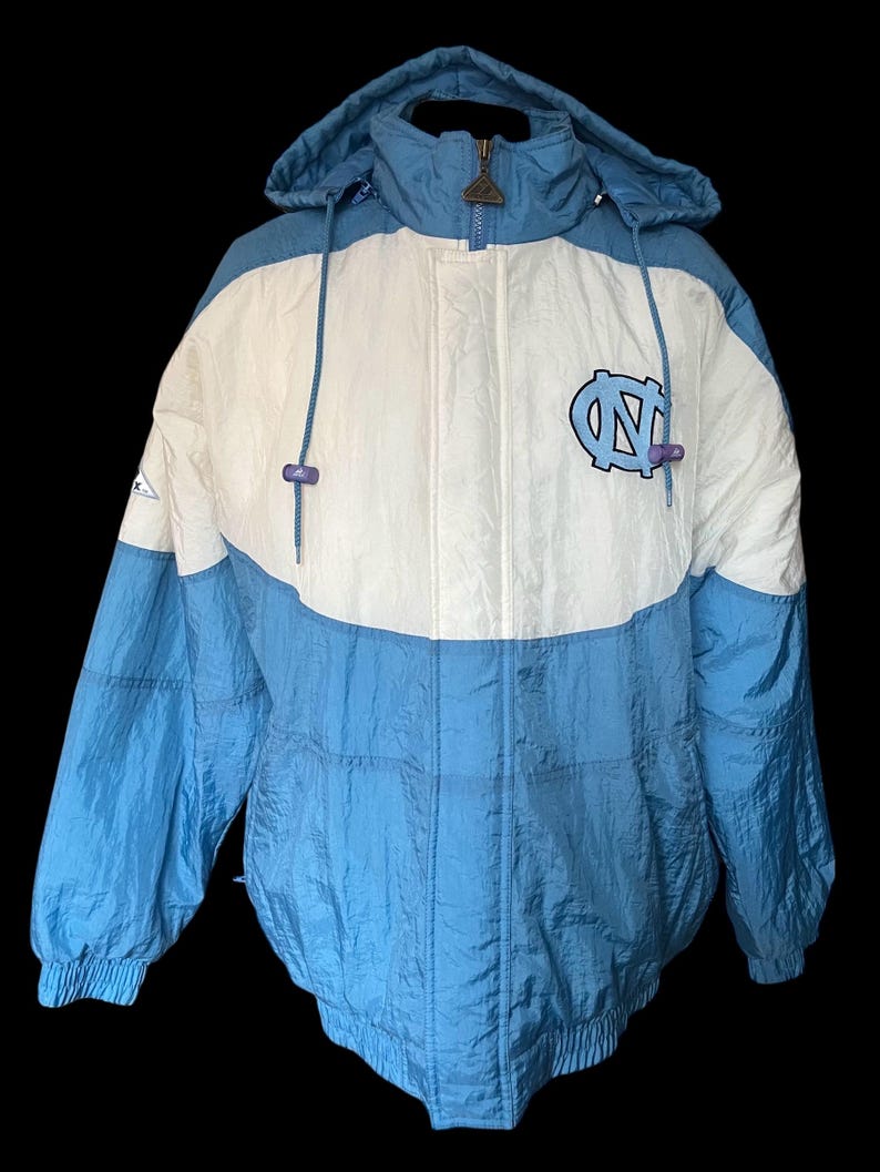 K&ouml;nnte beinhalten: Eine Kapuzenjacke in Hellblau und Wei&szlig; mit einem Colorblock-Design. Die Jacke hat einen Rei&szlig;verschluss und ein Logo auf der Brust. Die Jacke besteht aus einem gl&auml;nzenden, m&ouml;glicherweise Nylon-Material.