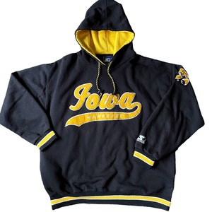以下が含まれることがあります： 黄色のアクセントが付いた黒いフード付きスウェットシャツ。スウェットシャツには、「Iowa」という単語が黄色のスクリプトで書かれており、その下に「Hawkeyes」と書いてあります。スウェットシャツは、裾と袖に黄色と黒のストライプのバンドが付いています。