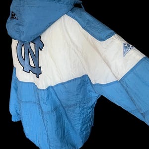 K&ouml;nnte beinhalten: Hellblaue und wei&szlig;e Kapuzenjacke mit dem Logo der University of North Carolina. Die Jacke hat ein Colorblock-Design mit einer hellblauen Kapuze und einem unteren Bereich sowie einem wei&szlig;en oberen Bereich. Das Logo befindet sich in der Mitte.