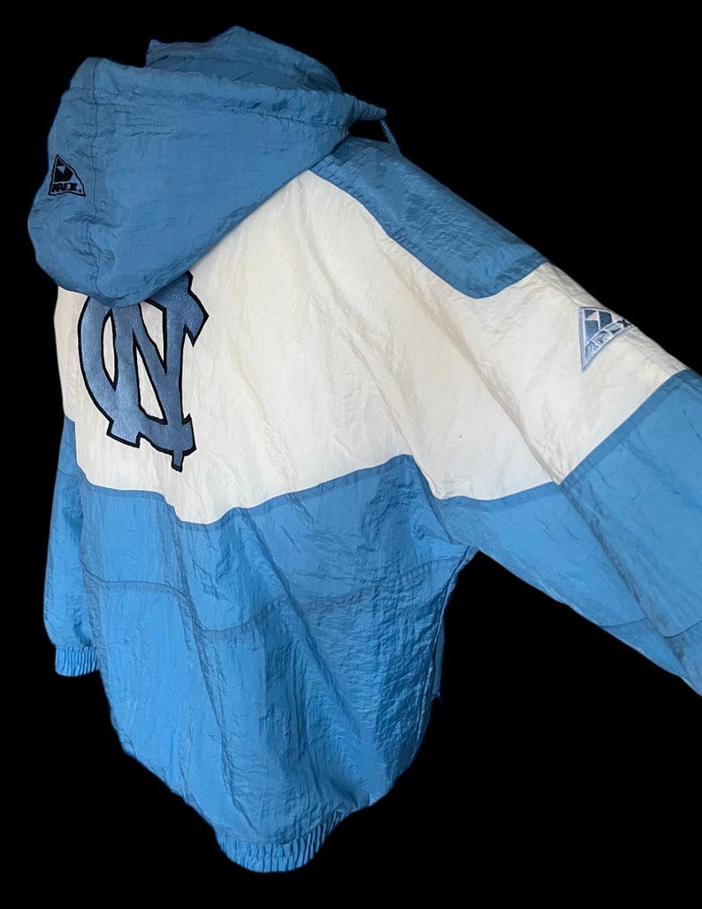 K&ouml;nnte beinhalten: Hellblaue und wei&szlig;e Kapuzenjacke mit dem Logo der University of North Carolina. Die Jacke hat ein Colorblock-Design mit einer hellblauen Kapuze und einem unteren Bereich sowie einem wei&szlig;en oberen Bereich. Das Logo befindet sich in der Mitte.