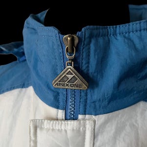 K&ouml;nnte beinhalten: Nahaufnahme einer blau, wei&szlig; und silberfarbenen Apex One Jacke. Die Jacke hat einen blauen Kragen und Rei&szlig;verschluss, mit dem Apex One Logo auf dem Rei&szlig;verschlussanh&auml;nger. Die Jacke ist aus einem leichten, m&ouml;glicherweise Nylon-Material.