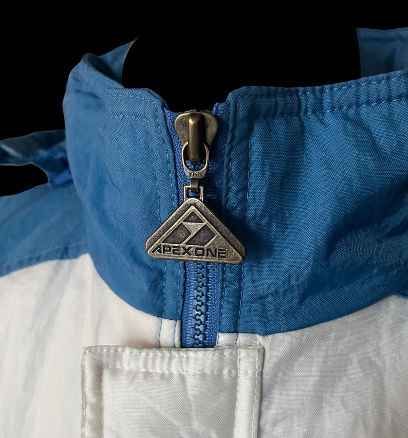 K&ouml;nnte beinhalten: Nahaufnahme einer blau, wei&szlig; und silberfarbenen Apex One Jacke. Die Jacke hat einen blauen Kragen und Rei&szlig;verschluss, mit dem Apex One Logo auf dem Rei&szlig;verschlussanh&auml;nger. Die Jacke ist aus einem leichten, m&ouml;glicherweise Nylon-Material.