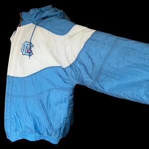 K&ouml;nnte beinhalten: Hellblaue und wei&szlig;e Nylonjacke mit Kapuze. Die Jacke hat ein Logo auf der Brust und einen Frontrei&szlig;verschluss. Die Jacke hat eine Vordertasche und einen Kordelzug an der Kapuze. Vintage-Stil.