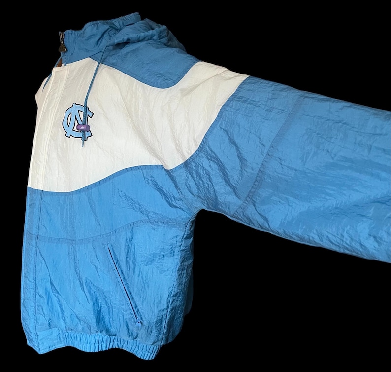 K&ouml;nnte beinhalten: Hellblaue und wei&szlig;e Nylonjacke mit Kapuze. Die Jacke hat ein Logo auf der Brust und einen Frontrei&szlig;verschluss. Die Jacke hat eine Vordertasche und einen Kordelzug an der Kapuze. Vintage-Stil.