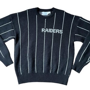 Könnte beinhalten: Schwarzer Pullover mit weißen vertikalen Streifen und dem Wort "RAIDERS" auf der Brust. Der Pullover hat einen gerippten Kragen, Bündchen und Saum. Die Ärmel haben ebenfalls weiße Streifen.