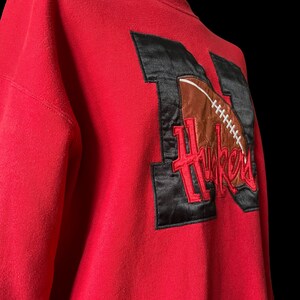 May include: Red sweatshirt with a black and brown embroidered patch featuring a football and the word "Huskers".