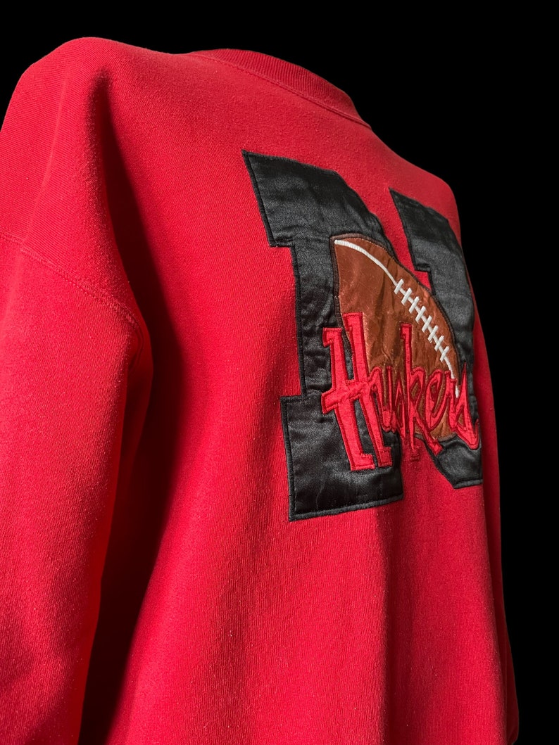 May include: Red sweatshirt with a black and brown embroidered patch featuring a football and the word "Huskers".