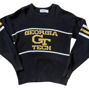 May include: Black knitted sweater featuring the words "GEORGIA TECH" in gold and white. The sweater has white stripes across the chest and on the sleeves, with gold accents. It has a crew neck and ribbed cuffs.
