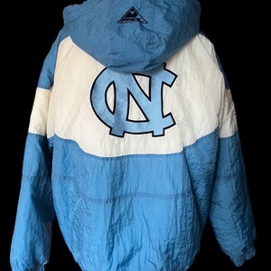 K&ouml;nnte beinhalten: Hellblaue und wei&szlig;e Kapuzenjacke mit gro&szlig;em "NC"-Logo auf dem R&uuml;cken. Die Jacke hat eine Kapuze mit Kordelzug, elastische B&uuml;ndchen und ein schwarzes Logo auf der Kapuze. Die Jacke ist aus leichtem Material, m&ouml;glicherweise Nylon.