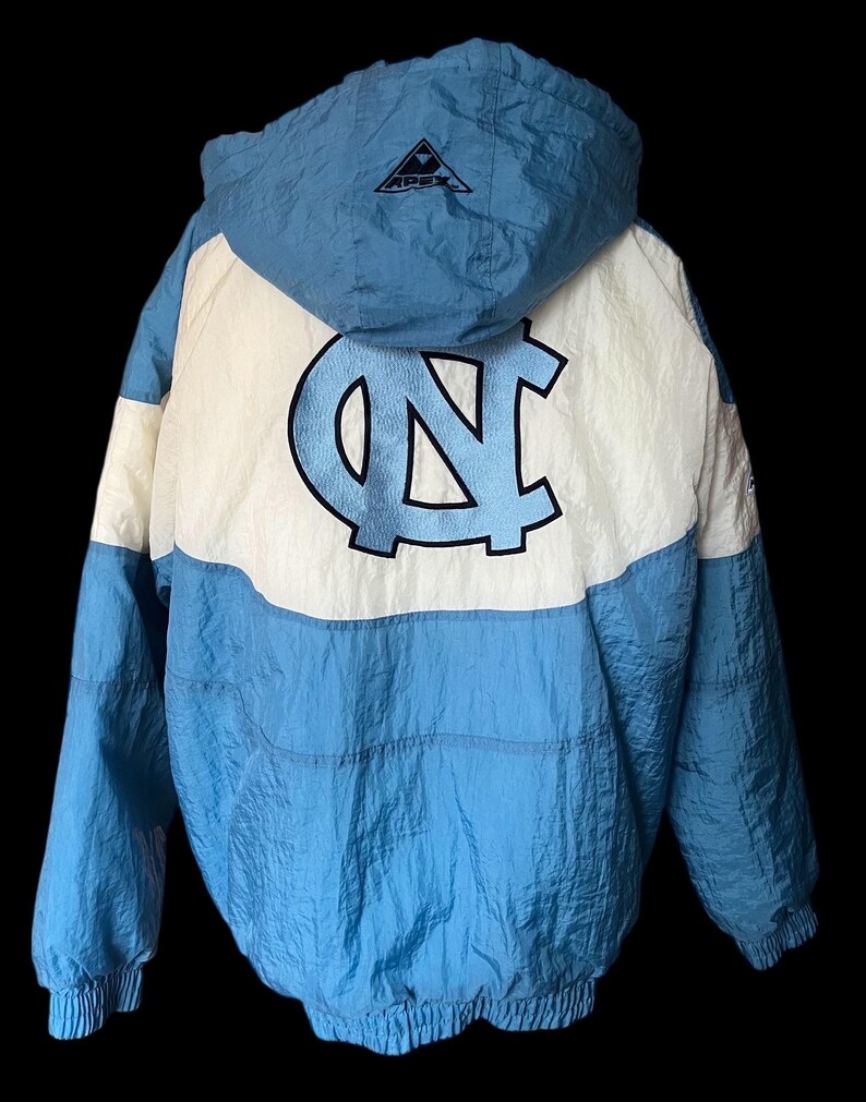 K&ouml;nnte beinhalten: Hellblaue und wei&szlig;e Kapuzenjacke mit gro&szlig;em "NC"-Logo auf dem R&uuml;cken. Die Jacke hat eine Kapuze mit Kordelzug, elastische B&uuml;ndchen und ein schwarzes Logo auf der Kapuze. Die Jacke ist aus leichtem Material, m&ouml;glicherweise Nylon.