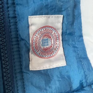K&ouml;nnte beinhalten: Nahaufnahme einer blau-wei&szlig;en Jacke mit Rei&szlig;verschluss. Ein wei&szlig;es Etikett mit einem rot-blauen kreisf&ouml;rmigen Logo mit der Aufschrift "LICENSED COLLEGIATE PRODUCTS OFFICIALLY" ist auf die Jacke gen&auml;ht.