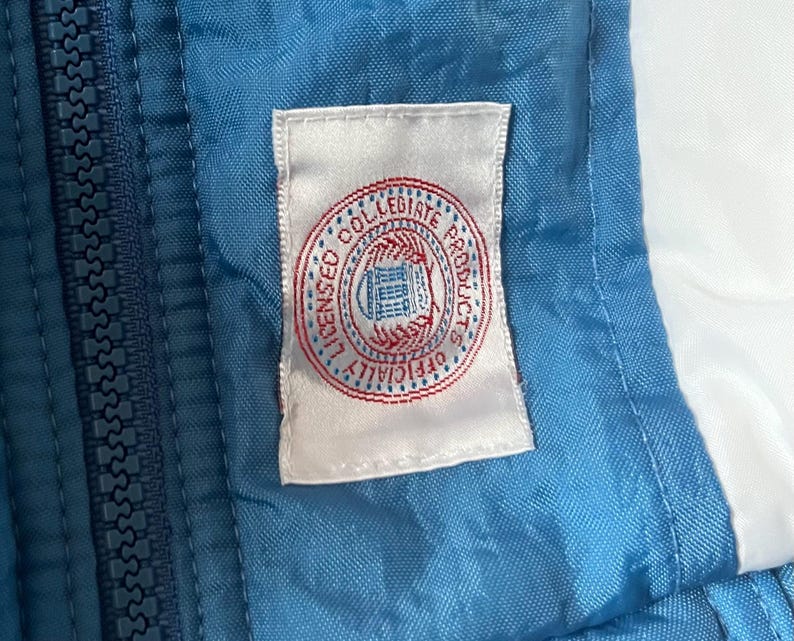 K&ouml;nnte beinhalten: Nahaufnahme einer blau-wei&szlig;en Jacke mit Rei&szlig;verschluss. Ein wei&szlig;es Etikett mit einem rot-blauen kreisf&ouml;rmigen Logo mit der Aufschrift "LICENSED COLLEGIATE PRODUCTS OFFICIALLY" ist auf die Jacke gen&auml;ht.