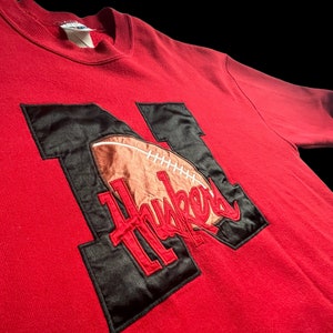 May include: Red sweatshirt with a black and brown embroidered football and the word "Huskers" on it.