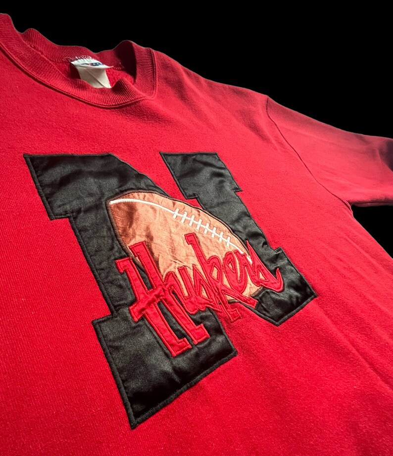May include: Red sweatshirt with a black and brown embroidered football and the word "Huskers" on it.
