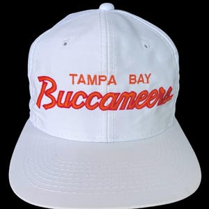 Könnte beinhalten: Weiße Baseballkappe mit den Worten "Tampa Bay Buccaneers" in Orange und Rot gestickt.
