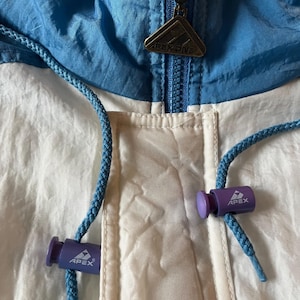 K&ouml;nnte beinhalten: Nahaufnahme einer Vintage Apex One Jacke. Die Jacke hat ein Colorblock-Design mit wei&szlig;en, hellblauen und dunkelblauen Eins&auml;tzen. Sie hat einen Rei&szlig;verschluss mit einem Apex One Metallanh&auml;nger und blaue Kordelz&uuml;ge mit lila Kordelstoppern.