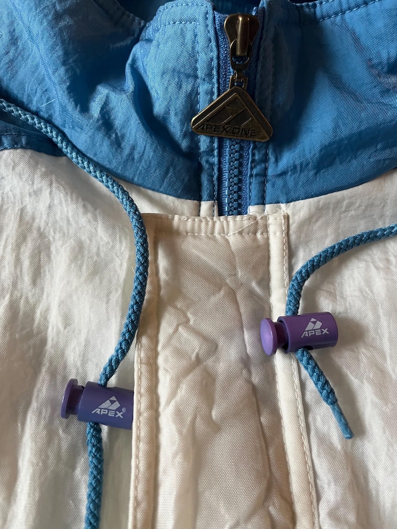 K&ouml;nnte beinhalten: Nahaufnahme einer Vintage Apex One Jacke. Die Jacke hat ein Colorblock-Design mit wei&szlig;en, hellblauen und dunkelblauen Eins&auml;tzen. Sie hat einen Rei&szlig;verschluss mit einem Apex One Metallanh&auml;nger und blaue Kordelz&uuml;ge mit lila Kordelstoppern.