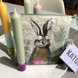 Könnte beinhalten: Eine Reißverschlusstasche mit einer Hasenillustration, zusammen mit drei konischen Kerzen in Gelb-, Grün- und Rosatönen. Die Tasche hat ein Etikett mit der Aufschrift "KATUI". Die Kerzen stehen auf einem weißen Tablett.