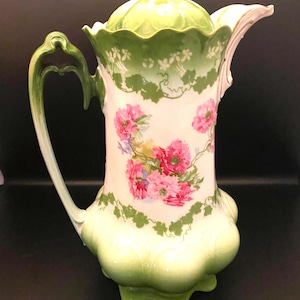 May include: A vintage porcelain coffee pot with a floral design. The pot features a light green lid and base, a white body adorned with pink flowers, and green vine accents. The handle and spout are also light green.