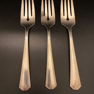 May include: Three silver forks with a decorative star-shaped cutout in the tines. The forks have a classic design with a tapered handle and a detailed, stylized end. The forks are arranged on a dark surface.