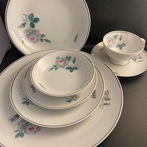May include: A set of white ceramic dinnerware with a delicate floral design featuring pink and white roses with green leaves. The set includes a large dinner plate, a salad plate, a bowl, a saucer, and a teacup.