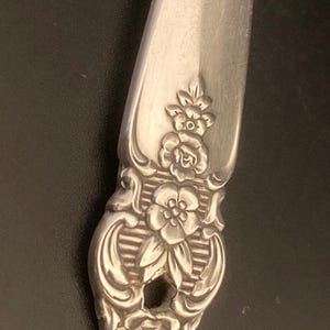 May include: Close-up of the ornate handle of a silver spoon with a floral design. The handle is decorated with three roses and a smaller flower in the center.