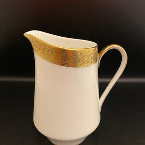 May include: White ceramic creamer with a gold rim and intricate gold floral pattern. The creamer has a rounded body and a handle.