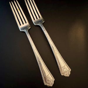 May include: Two silver forks with ornate designs on the handles. The forks are laid out parallel to each other.