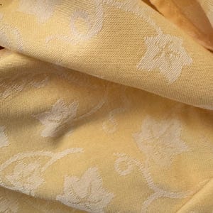 May include: Close-up of a yellow fabric with a repeating pattern of white floral and vine-like designs. The fabric is folded, showing texture and depth. The color is a soft, warm yellow.