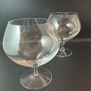 May include: Two clear glass brandy snifters with a round bowl and a tall stem. The glasses are empty and have a slight swirl pattern.