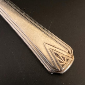 May include: Close-up of the handle of a silver fork with an Art Deco design. The handle is decorated with a geometric pattern of triangles and lines.