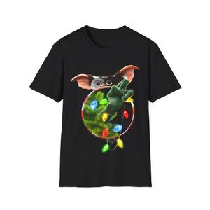 May include: Black t-shirt with a festive Gremlin design. The graphic includes a Gremlin character, a Christmas tree, and colourful string lights. The Gremlin is giving a peace sign with a green hand, inside a red and white circle.